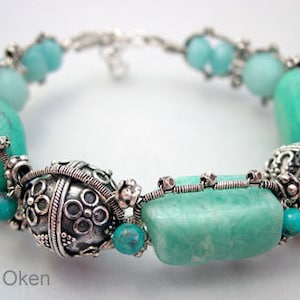 Op de afbeelding: Een zilveren armband met turquoise en groene stenen. De armband heeft een verscheidenheid aan kralen, waaronder ronde, ovale en rechthoekige vormen. De stenen zijn ingelegd in zilverdraad en zijn versierd met zilveren kralen.