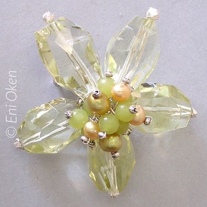 Puede incluir: Un broche con forma de flor hecha de vidrio facetado transparente con un grupo de cuentas verdes y doradas en el centro.