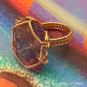 Peut inclure: Une bague en fil d'or avec une grande améthyste violette facetée. La bague est placée sur un fond coloré.