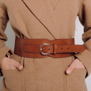 May include: A brown leather belt with a gold buckle. The belt is wide and has a curved shape. It is worn over a brown coat.