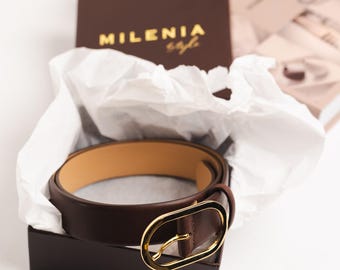 Peut inclure: Une ceinture en cuir marron avec une boucle ovale dorée. La ceinture est dans une boîte marron avec le texte "MILENIA style" sur le dessus.