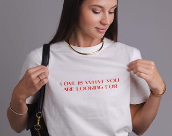 Könnte beinhalten: Weißes T-Shirt mit rotem Text "LOVE IS WHAT YOU ARE LOOKING FOR". Dazu blaue Jeans, ein schwarzer Gürtel und eine schwarze Handtasche. Eine goldene Halskette und ein Armband ergänzen den Look.