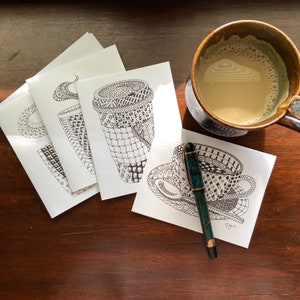 May include: Four white cards with black and white illustrations of coffee cups. The cards are arranged in a fan shape. A black and green pen is lying on top of the cards. A brown ceramic mug with a beige interior is in the background.