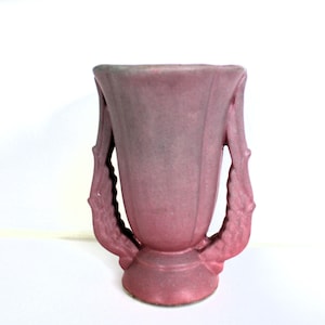 May include: A pink ceramic vase with two handles in the shape of stylized leaves. The vase has a textured, slightly iridescent finish.