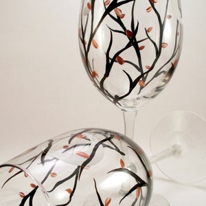 May include: Two clear wine glasses with a hand-painted design. The design features black branches with small, orange-red leaves. One glass is upright, the other is lying on its side. The glasses are on a white background.
