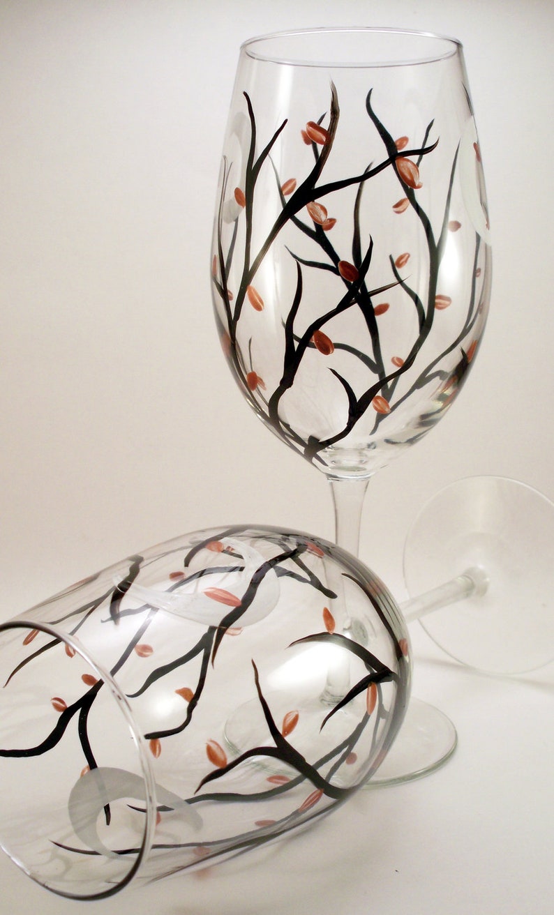 May include: Two clear wine glasses with a hand-painted design. The design features black branches with small, orange-red leaves. One glass is upright, the other is lying on its side. The glasses are on a white background.
