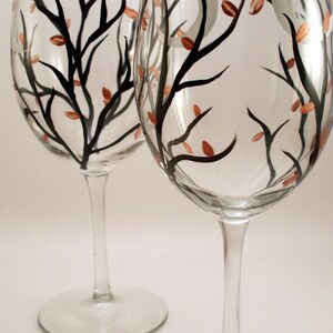May include: Two clear wine glasses with a hand-painted design. The design features black tree branches with small copper-colored leaves and a white crescent moon. The glasses have a tall stem and a round base.