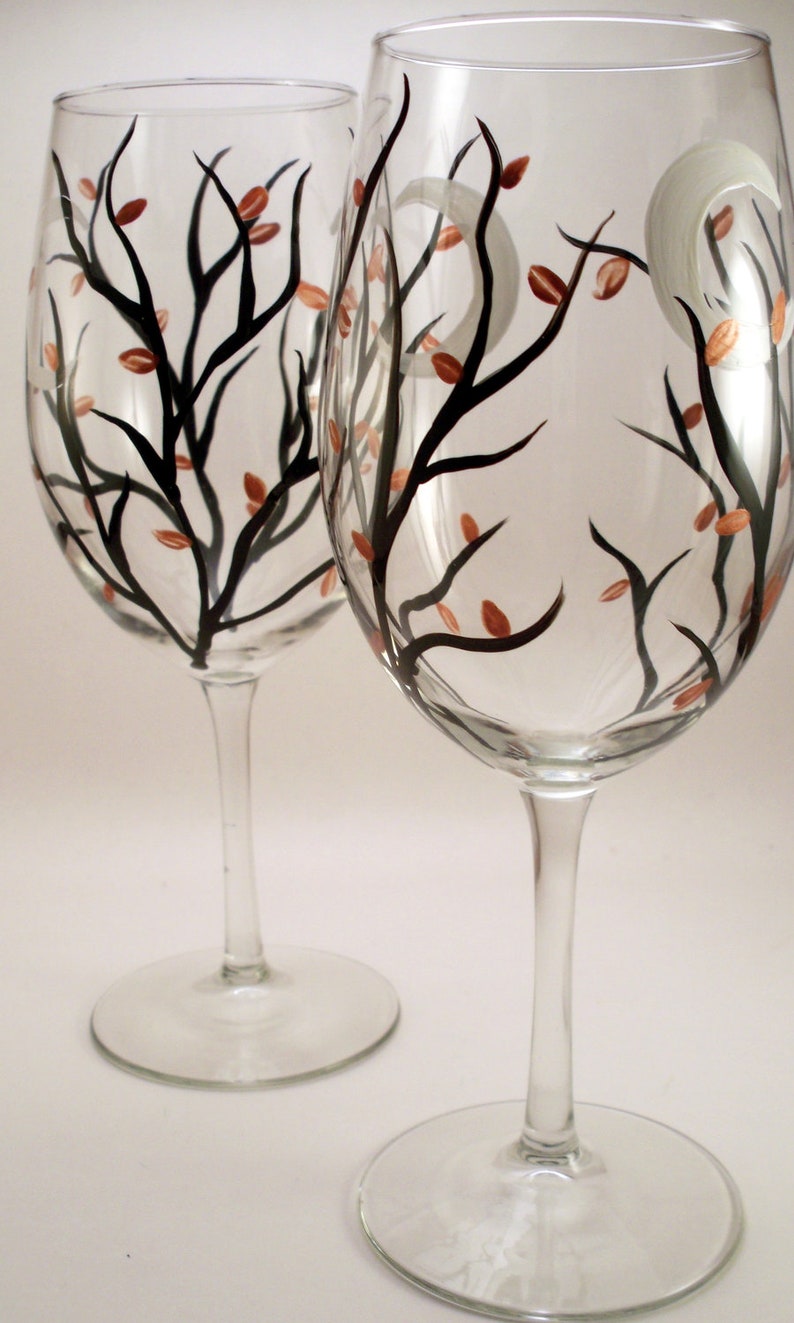 May include: Two clear wine glasses with a hand-painted design. The design features black tree branches with small copper-colored leaves and a white crescent moon. The glasses have a tall stem and a round base.