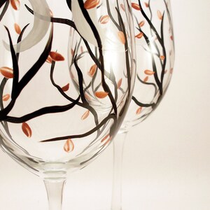May include: Two clear wine glasses with hand-painted designs. The glasses feature black branches, orange leaves, and a gray crescent moon. The stems and bases are clear, and the background is white.