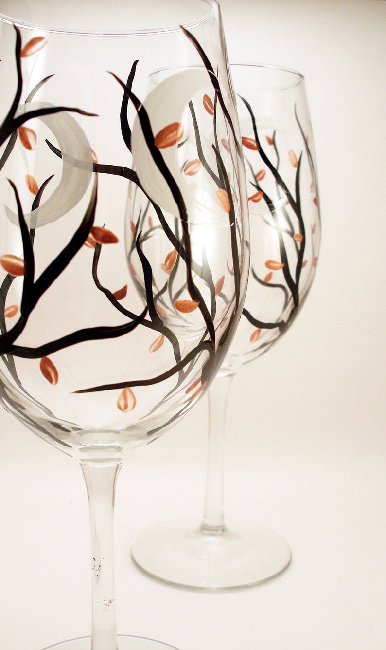 May include: Two clear wine glasses with hand-painted designs. The glasses feature black branches, orange leaves, and a gray crescent moon. The stems and bases are clear, and the background is white.