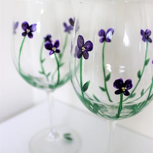 May include: Two clear glass wine glasses with hand-painted purple flowers and green stems. The flowers have yellow centers.
