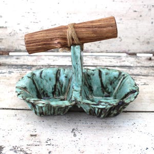 May include: A handcrafted ceramic serving dish with two compartments, glazed in a turquoise and black mottled pattern. It features a wooden handle secured with twine, perfect for serving dips or snacks.