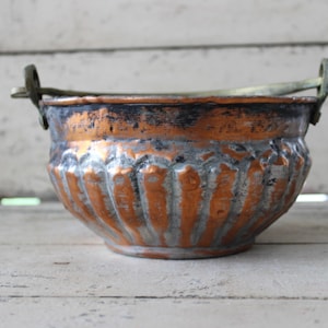 May include: Antique copper bowl with a decorative, fluted design. The bowl has a dark patina and a brass handle. The copper bowl is a vintage piece, perfect for home decor or as a unique kitchen accent.