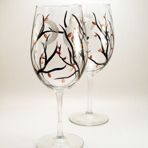 May include: Two clear wine glasses with a hand-painted design. The design features black branches, copper-colored leaves, and white crescent moons. The glasses have a tall stem and a wide bowl.