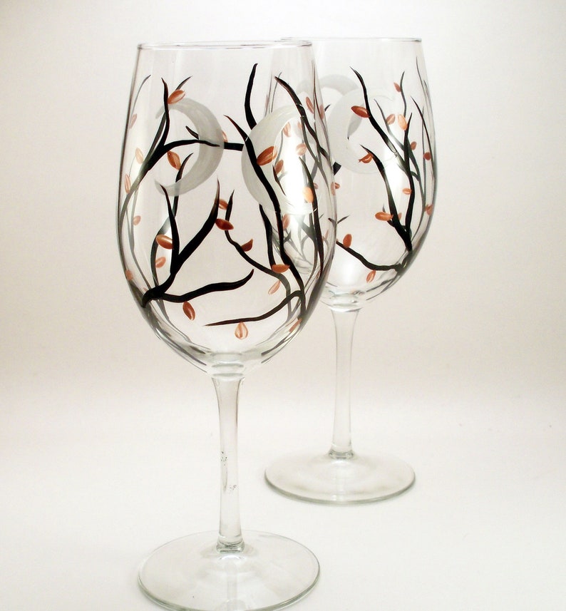 May include: Two clear wine glasses with a hand-painted design. The design features black branches, copper-colored leaves, and white crescent moons. The glasses have a tall stem and a wide bowl.