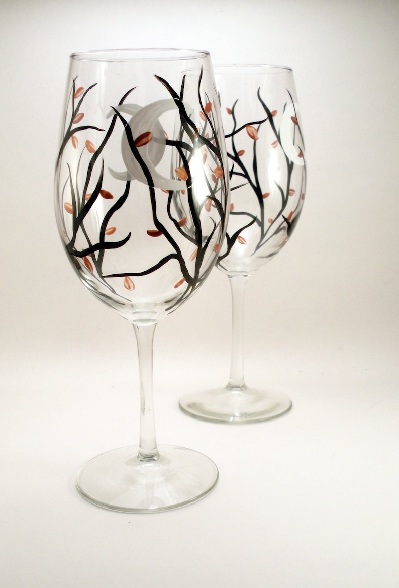 May include: Two clear wine glasses with a hand-painted design. The design features black branches, small orange leaves, and a white crescent moon. The glasses have a classic stem and base.