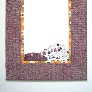 May include: A rectangular picture frame with a patterned, dark purple fabric exterior. The frame's interior edge features a colorful, patterned border. The frame's interior is white, with a decorative fabric element at the bottom.
