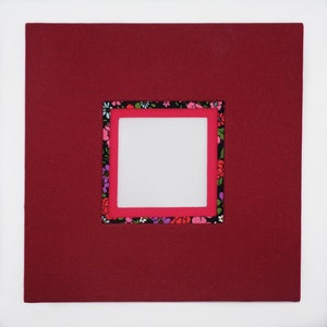 May include: A square, burgundy-colored mat with a central square opening. Inside the opening is a smaller square, framed in bright pink, and surrounded by a floral patterned fabric. The overall design is a layered, framed aesthetic.