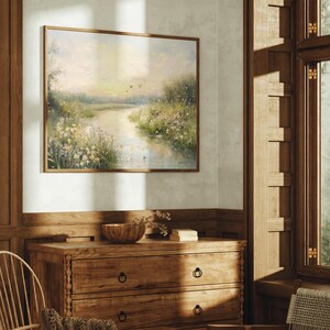 May include: A framed painting of a landscape with a river winding through a field of wildflowers. The painting is in a light brown frame and hangs on a white wall.