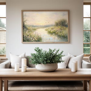 May include: A living room interior with a light wood coffee table, a white sofa with two white pillows, a potted green plant, and a framed painting of a river scene with a sunset in the background.