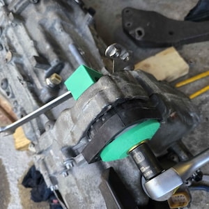 May include: A green plastic tool is used to tighten a bolt on a car transmission. The bolt is being tightened with a wrench. The wrench has a 19mm socket.