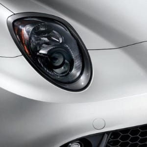 May include: Close-up of a white car's front end, showcasing a sleek headlight design. The headlight has a dark interior with a chrome trim, and an orange side marker. The car's body has smooth curves and a black grill.