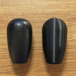 May include: Two black, teardrop-shaped objects sit side-by-side on a wood-grain surface. The object on the left has a textured surface, while the one on the right has a smooth, ridged surface. The objects appear to be automotive parts.