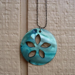 May include: A round, turquoise pendant necklace with a cut-out floral design. The pendant is suspended from a bronze-colored ball chain. The necklace is displayed against a light brown, textured background.