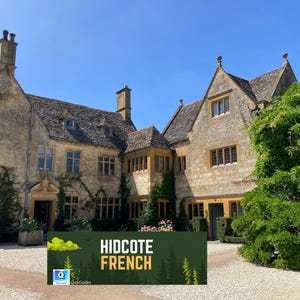 May include: A large, historic stone building with multiple gabled roofs and chimneys under a clear blue sky. The building has many windows and is partially covered in climbing plants. A sign in front reads "HIDCOTE FRENCH".