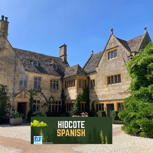 May include: A large, stone building with multiple gabled roofs and chimneys under a clear blue sky. The building is covered in climbing plants. A banner in front reads "HIDCOTE SPANISH".
