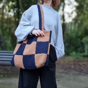 Peut inclure: Un grand sac fourre-tout à carreaux bleu marine et beige. Le sac a une bandoulière assortie et est porté à l'épaule. La personne porte un sweat-shirt bleu clair et un pantalon noir. Le sac est en tissu doux.