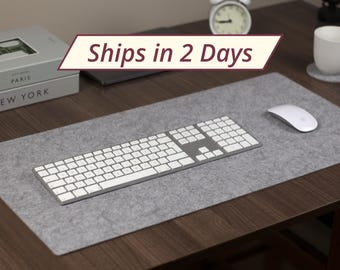 Könnte beinhalten: Eine graue Filz-Schreibtischunterlage mit einer weißen Tastatur und einer weißen Maus. Die Schreibtischunterlage liegt auf einem Holztisch.