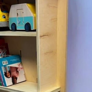 May include: A light-colored wooden children's desk and bookshelf unit. The unit has two shelves with toys and books. A book titled "Doctor" is on the desk. A small wooden chair is in front of the desk.