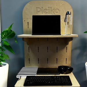 May include: A light wood standing desk with a black laptop, a black keyboard, a black and white striped notebook, a black tablet, and black headphones. The desk has a pegboard with holes and the text "Pleiko" is engraved on the top shelf.