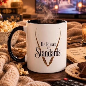 Op de afbeelding: Een witte keramische mok met een zwarte binnenkant en handvat. De voorzijde bevat de tekst "He Ruined my Standards" in zwarte letters, omlijst door twee gestileerde, puntige, bruine en gouden veerachtige afbeeldingen die samenkomen bij een rode edelsteen.