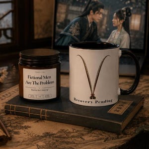 May include: A brown glass candle jar with a white label reading Fictional Men Are The Problem and a white ceramic mug with a black interior and handle featuring a V-shaped graphic and the text Recovery Pending, both resting on a dark book.