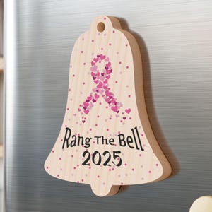 May include: Wooden bell-shaped magnet with a pink ribbon made of hearts and the text "Rang The Bell 2025". The bell is decorated with pink polka dots and has a hole at the top.