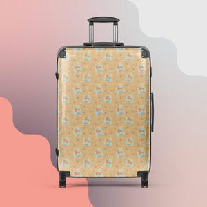 May include: A tan rolling suitcase featuring a repeating pattern of a cartoon horse, clouds, and the word "FRIDA" in blue. The suitcase has a retractable handle and black trim.