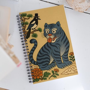 May include: Spiral-bound notebook featuring a blue tiger illustration with orange accents and a magpie wearing a top hat. The cover has a tan background with a black and white spiral binding.