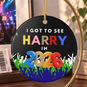 May include: A round black ceramic ornament featuring the white text I GOT TO SEE HARRY IN, with the colorful 3D-style numbers 2026 below. The bottom edge displays a white and blue silhouette graphic of a crowd with raised arms and hands.