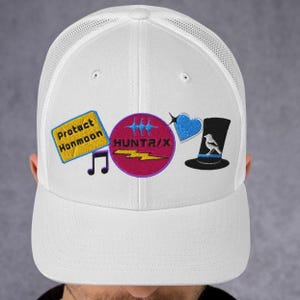 May include: White trucker hat with embroidered patches. Patches include text "Protect Honmoon", a musical note, a circle with "HUNTRIX" and lightning bolt, a blue heart, and a black top hat with a bird.