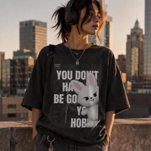 May include: Dark gray t-shirt with the text "YOU DON'T HAVE TO BE GOOD AT YOUR HOBBY" and a white bunny graphic. The shirt also has the text "STRAY KIDS" on the upper left.