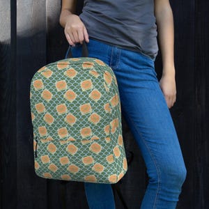 Può includere: Zaino verde con un motivo ripetuto di quadrati arancioni e un design a scaglie bianche. Lo zaino ha un manico nero e una tasca frontale. La borsa è tenuta da una persona che indossa una maglietta grigia e jeans blu.