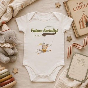 Könnte beinhalten: Ein weißer kurzärmeliger Babybody mit einer Grafik eines Affen am Trapez. Der Text lautet "Born to Fly Future Aerialist Est. 2025". Das Design enthält ein leeres weißes Banner, das vom Affen gehalten wird, sowie gedeckte Erdtöne.