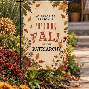 May include: A rectangular garden flag featuring a cream-colored background with a border of autumn leaves and flowers. The text reads: MY FAVORITE SEASON IS THE FALL OF THE PATRIARCHY. The flag is suspended from a black metal garden stake.