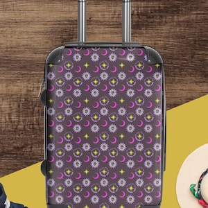 May include: A patterned suitcase featuring yellow stars, pink moons, and white floral designs on a dark purple backdrop. The case has a retractable handle and black trim. The suitcase is designed for travel.