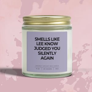 May include: A glass jar candle with a gold lid and a purple label. The label reads "SMELLS LIKE LEE KNOW JUDGED YOU SILENTLY AGAIN." The candle is 4 oz and burns for 20 hours. The candle is made of coconut apricot wax.