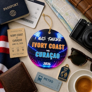 May include: A round ceramic ornament featuring a stadium background with bright lights and the text I WAS THERE IVORY COAST VS CURAÇAO 2026. The design includes orange and light blue lettering, suspended by a thin gold string for display.