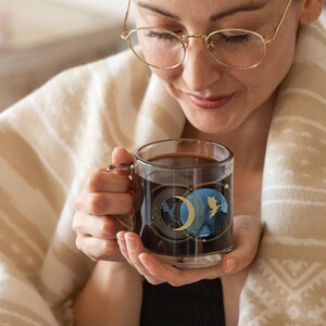 May include: A clear glass mug filled with dark liquid, featuring a celestial design with a crescent moon and a blue and gold planet. The mug is held by a person wrapped in a beige blanket.