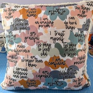 May include: Square cushion with a white base, featuring colorful cloud shapes filled with positive affirmations in black script. The cushion has a fluffy, cream-coloured trim.